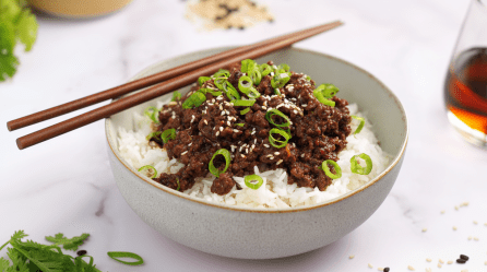 Eine Schale Bulgogi mit Hackfleisch auf hellem Untergrund. Auf der Schale liegen zwei Essstäbchen. Der Gericht ist mit Frühlingszwiebeln und Sesamsamen garniert.