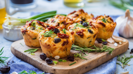 Sechs Focaccia Muffins stehen auf einem Holzbrettchen. Um sie herum verteilt liegen einige schwarze Oliven, Rosmarinzweige und Frühlingszwiebelringe. Im Hintergrund sieht man noch Frühlingszwiebeln, eine Flasche mit Öl und eine Knoblauchknolle.