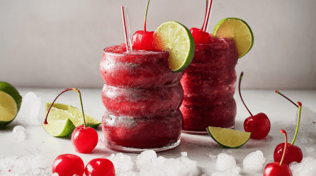 Zwei Gläser mit Frozen Cherry Cola stehen auf einem weißen Untergrund vor einem weißen Hintergrund. Sie sind mit Kirschen und Limettenscheiben garniert. Vor den Gläsern liegen Kirschen, Limetten sowie crushed ice.