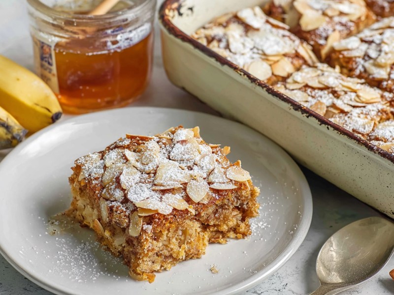 Ein Teller mit einem Stück der gebackenen Mandelcroissant-Haferflocken. Daneben steht die Auflaufform voll mit den Baked Oats. Außerdem sind Bananen, einzelne Mandeln, ein Löffel und ein Glas Honig im Bild zu sehen.