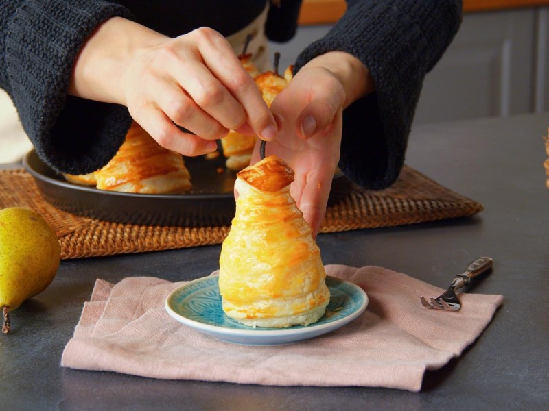 Eine gefüllte Birne im Blätterteig-Mantel, die von Köchin Adrianna auf einem kleinen Teller platziert wird. Im Bild sind noch eine frische Birne auf der Arbeitsfläche, eine weitere gefüllte Birne. Im rechten Bildrand steht ein Korb.