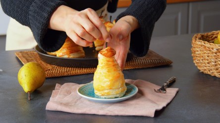 Eine gefüllte Birne im Blätterteig-Mantel, die von Köchin Adrianna auf einem kleinen Teller platziert wird. Im Bild sind noch eine frische Birne auf der Arbeitsfläche, eine weitere gefüllte Birne. Im rechten Bildrand steht ein Korb.