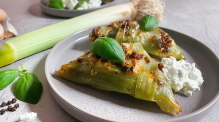 Ein Teller mit zwei dreieckig geformten gefüllten Lauchblättern, die mit einem Blatt Basilikum garniert sind. Auf dem Teller sind sie mit einem Dip angerichtet. Im Hintergrund liegt ein ganzer Lauch. Zudem liegen einige ganze Pfefferkörner und Knoblauchzehen herum.