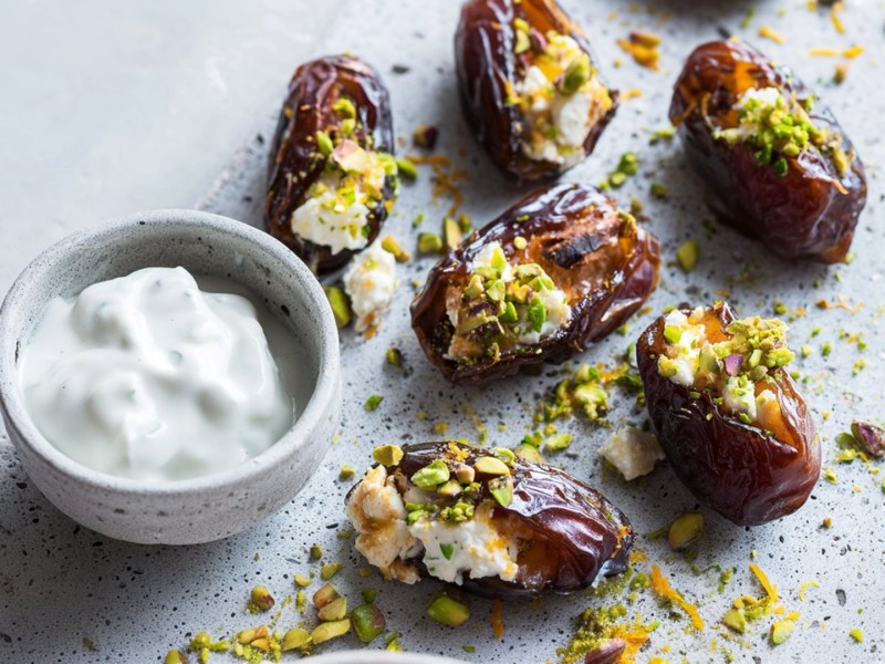 Ein grauer Steinteller, auf dem einige gefüllte Datteln liegen, die zuvor gegrillte Dattel-Feta-Spieße waren. Die Datteln werden mit einer kleinen Schale Joghurt-Dip serviert.