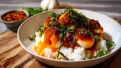 Eine Schüssel mit Reis und Gochujang-Eiern. Das Ganze ist mit Nori und Frühlingszwiebeln garniert. Im Hintergrund kann man eine Schale Chiliflocken sowie eine Knoblauchknolle erkennen.