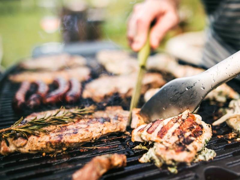 Ein Grill, auf dem einiges an Grillfleisch liegt. Man kann angedeutet jemand erkennen, der das Grillfleisch mit Grillwerkzeug wendet.