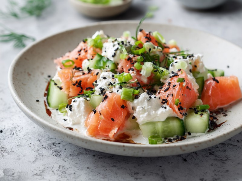 Ein Teller mit Gurkensalat mit Hüttenkäse und Lachs. Das Ganze ist mit schwarzem Sesam, Frühlingszwiebeln und Dill garniert.