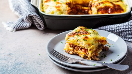 Ein Stück Gyros-Kartoffel-Auflauf liegt vor einer Auflaufform mit noch mehr Auflauf darin.