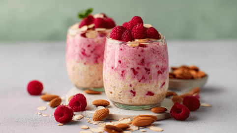 Zwei Gläser mit Himbeer-Mandel-Overnight-Oats, darum liegen Früchte und Mandeln als Dekoration