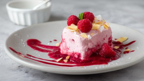 Ein Himbeer-Parfait, dass auf etwas Himbeerpüree angerichtet ist und mit frischen Himbeeren, zerkrümeltem Baiser und gerösteten Mandelblättchen garniert ist.