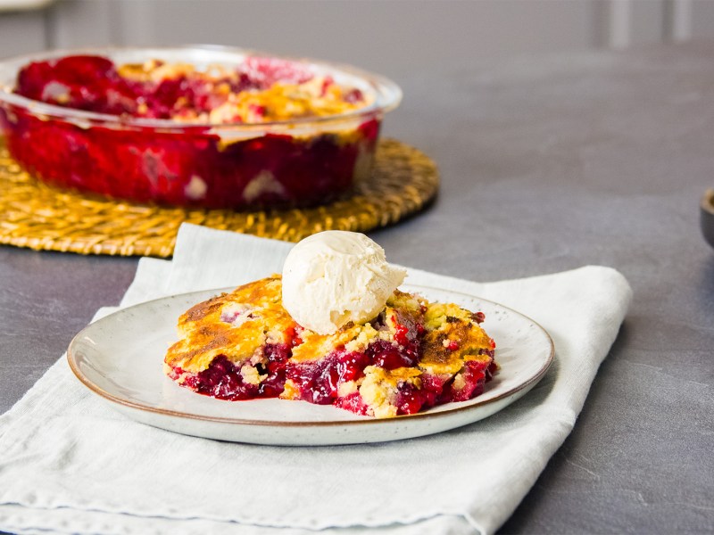 Ein Stück Himbeerkuchen-Auflauf mit einer Kugel Vanilleeis auf einem Teller. Im Hintergrund steht eine Auflaufform mit dem Auflauf.