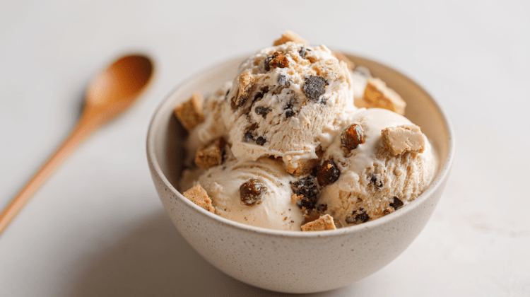 Eine Schale Keksteig-Eis. Links im Hintergrund liegt ein Holzlöffel. Das Eis ist mit Keksteig-Stücken, Schokodrops und karamellisierten Haselnüssen zubereitet.