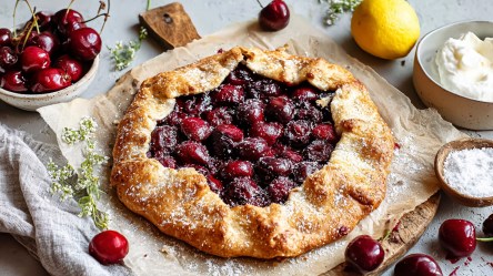 Eine Kirsch-Galette, die mit Puderzucker bestäubt ist. Drum herum liegen eine Schale mit Kirschen und eine mit frisch geschlagener Sahne und einzelne Kirschen sowie eine Zitrone verteilt. Auch eine kleine Schale mit Puderzucker ist zu sehen. Die Galette liegt auf einem rustikalen Brett, das mit Backpapier ausgelegt ist.