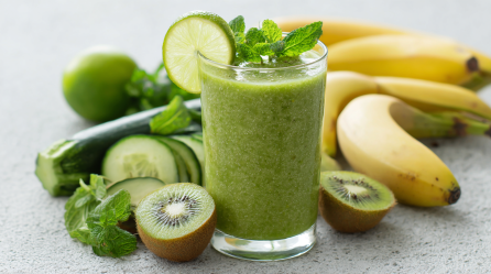 Ein Glas Kiwi-Zucchini-Smoothie auf einer grauen Arbeitsplatte. Um das Glas herum verteilt liegen Kiwis, Gurken, Bananen und eine Zucchini. Der Smoothie ist mit einer Limettenscheibe garniert.