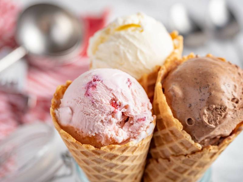 Drei Eiswaffeln mit jeweils einer Kugel Eis darin. Dabei handelt es sich um Erdbeer-, Vanille und Schokoladeneis, das mit der KLAMER 2-in-1 Eismaschine hergestellt wurde. Im Hintergrund kann man einen Eisportionierer sowie 2 Löffel sehen. Der Portionierer liegt auf einem rot-weißen Küchenhandtuch.