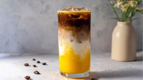Ein großes Glas mit Mango Iced Latte, das vor einem hellen minimalistischen Hintergrund platziert ist. Es liegen ein paar Kaffeebohnen verstreut. Im Hintergrund steht eine beige Vase mit Kamillen.