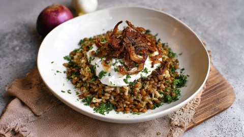 Ein Teller mit einer großen Portion Mujaddara, also Reis mit Linsen und karamellisiertem Zwiebeln. Alles ist mit etwas frischem Joghurt und frischen gehackten Kräutern. Im Hintergrund liegt ein rote Zwiebel und eine Knoblauchknolle.
