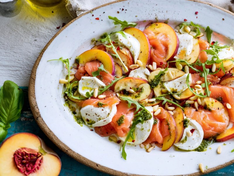 Ein großer Servierteller mit Nektarinen-Räucherlachs-Caprese. Alles ist mit Pinienkernen, frischem Rucola und etwas Basilikum-Pesto angerichtet. Um den Teller herum liegen eine halbe Nektarine, etwas frischer Basilikum und man kann angeschnitten Essig- und Ölgefäße erkennen.