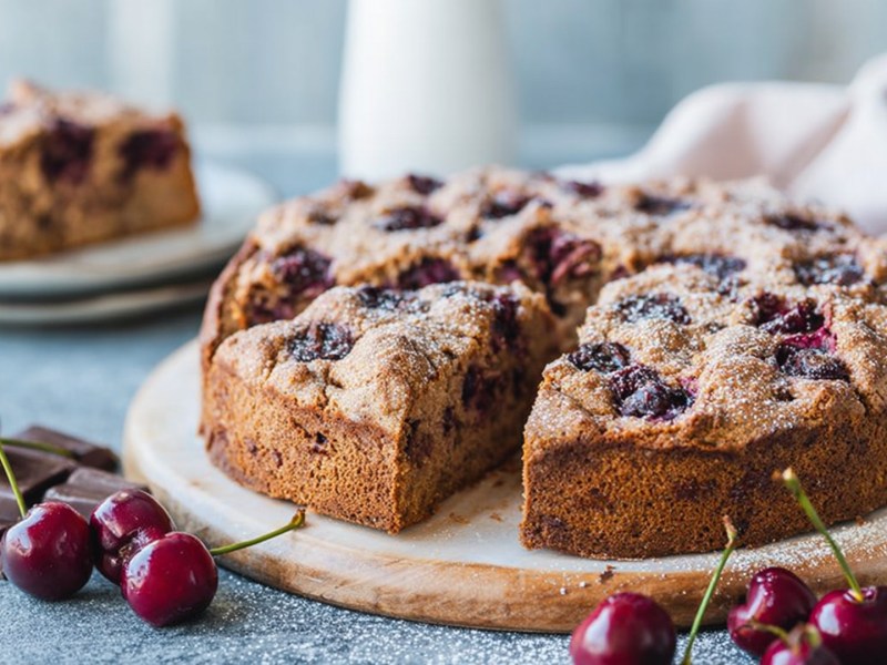 Ein rundes Holzbrett, auf dem ein runder angeschnittener Nusskuchen mit Kirschen und Schokolade platziert ist. Ringsum liegen frische Kirschen und einige Stücke Schokolade. Im Hintergrund steht ein Teller mit einem Stück Kuchen und ein Küchentuch liegt drapiert da.