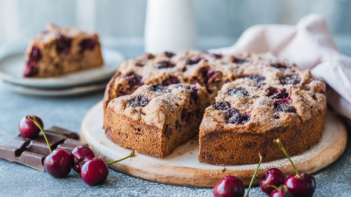 Ein rundes Holzbrett, auf dem ein runder angeschnittener Nusskuchen mit Kirschen und Schokolade platziert ist. Ringsum liegen frische Kirschen und einige Stücke Schokolade. Im Hintergrund steht ein Teller mit einem Stück Kuchen und ein Küchentuch liegt drapiert da.