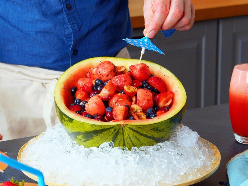 Ein Obstsalat aus der Wassermelonenschale. Die Schale ist in einer großen Schüssel mit Eiswürfeln platziert, damit der Salat schön kühl bleibt. Man kann Koch Oli erkennen, wie er ein Cocktailschirmchen in den Salat steckt. Rechts neben der Schüssel steht ein Glas mit einem Wassermelonencocktail.