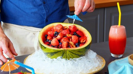 Ein Obstsalat aus der Wassermelonenschale. Die Schale ist in einer großen Schüssel mit Eiswürfeln platziert, damit der Salat schön kühl bleibt. Man kann Koch Oli erkennen, wie er ein Cocktailschirmchen in den Salat steckt. Rechts neben der Schüssel steht ein Glas mit einem Wassermelonencocktail.