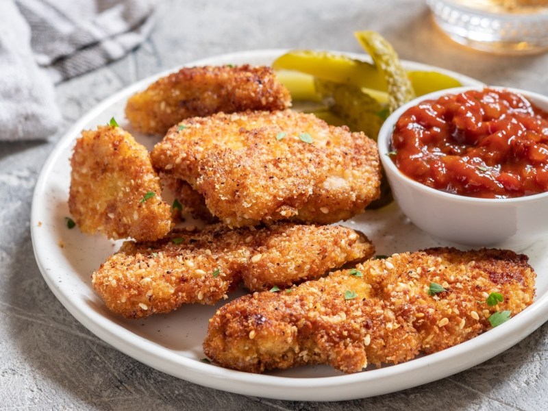 Ein Teller mit paniertem Kokos-Hähnchen, Gewürzgurken und einem Salsa-Dip.