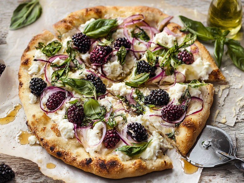 Eine Pizza mit Brombeeren und Ricotta, die mit frischem Basilikum garniert und zusätzlich mit roten Zwiebeln belegt ist. Die Pizza liegt auf Backpapier und ist mit Honig beträufelt. Daneben stehen eine Schale mit Brombeeren und eine mit geriebenem Käse. Außerdem ist ein Pizzaschneider zu erkennen.