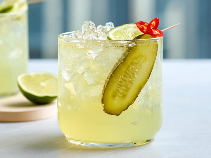 Ein Glas mit scharfer Essiggurken-Limonade, die mit einer langen Scheibe Essiggurke, einem Limettenstück und feinen Chiliringen garniert ist. Im Hintergrund ist ein weiteres Glas zu sehen, das auf einem Brett steht, auf dem auch eine angeschnittene Limette liegt.