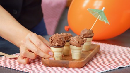Ein kleines Holzbrett, das auf einem rotgestreiften Tischuntersetzer steht. Auf dem Brett stehen einige Waffeln mit selbstgemachtem Schokoladen-Eis. In einer Eiskugel steckt ein kleines Cocktailschirmchen. Ein Hand greift nach einer der Eiswaffeln. Im Hintergrund kann man einen orangen Ballon erkennen.