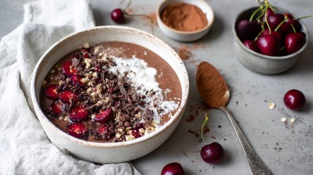 Eine Schwarzwälder Kirsch-Smoothie-Bowl, die mit frischen Kirschen, Granola, geraspelter dunkler Schokolade und Joghurt garniert ist. Neben der Schüssel ist ein helles Küchenhandtuch drapiert. Außerdem sind ein Löffel mit Kakopulver, eine Schale mit Kakao und eine Schale mit Kirschen zu sehen. Frische Kirschen kullern auch herum.