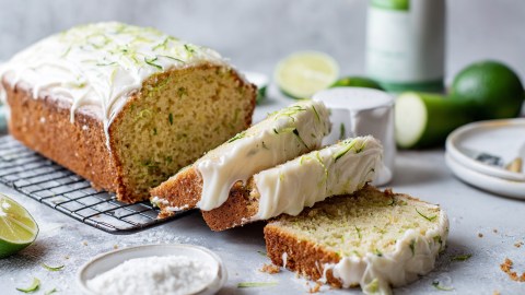 Ein aufgeschnittener süßer Zucchini-Kuchen mit einem weißen Frischkäse-Frosting. Ringsum liegen Zutaten wie frische Limetten, eine Schale mit Puderzucker und eine angeschnittene Zucchini herum sowie weitere Gefäße und ein Teller.