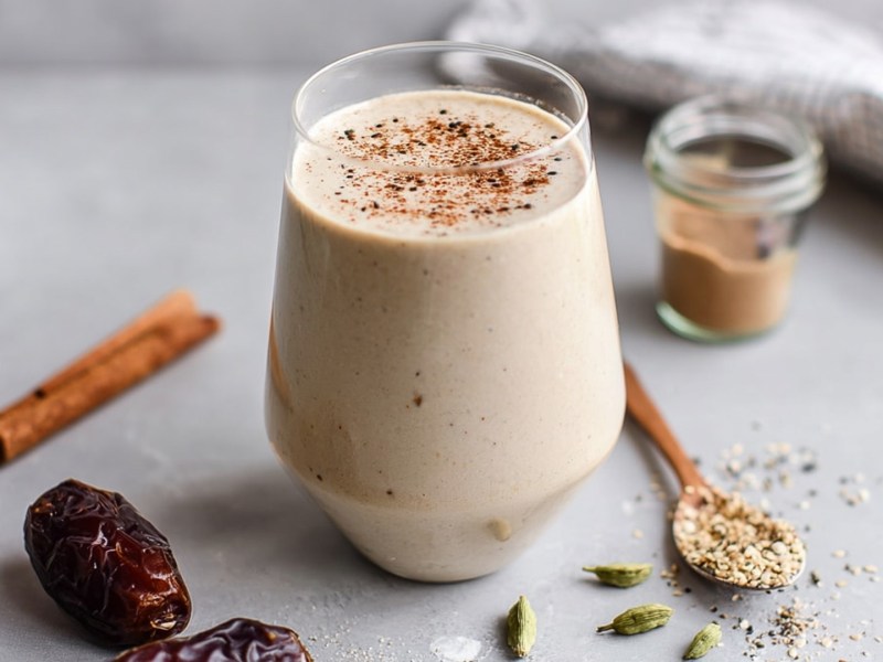 Ein Glas mit Tahini-Dattel-Smoothie, das mit etwas Zimt garniert ist. Ringsum das Glas liegen Zutaten verstreut wie Datteln, eine Zimtstange, ein Löffel mit Sesam und ein kleines Glas mit Sesam-Mus.