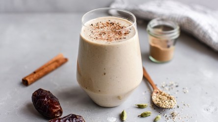Ein Glas mit Tahini-Dattel-Smoothie, das mit etwas Zimt garniert ist. Ringsum das Glas liegen Zutaten verstreut wie Datteln, eine Zimtstange, ein Löffel mit Sesam und ein kleines Glas mit Sesam-Mus.
