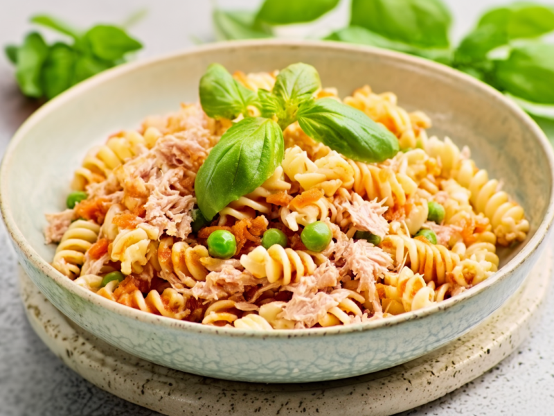 Thunfisch-Pasta in einem Teller, garniert mit frischem Basilikum. Im Hintergrund liegt frischer Basilikum als Deko.