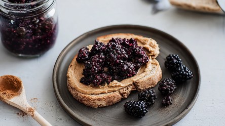 Ein Toast mit Mandelmus und Brombeeren, der auf einem dunklen rustikalen Teller liegt. Auf dem Teller liegen noch einige einzelne Beeren. Neben dem Teller liegt ein Holzlöffel mit Zimt. Im Hintergrund kann man ein Glasgefäß mit frischen Brombeeren sehen.