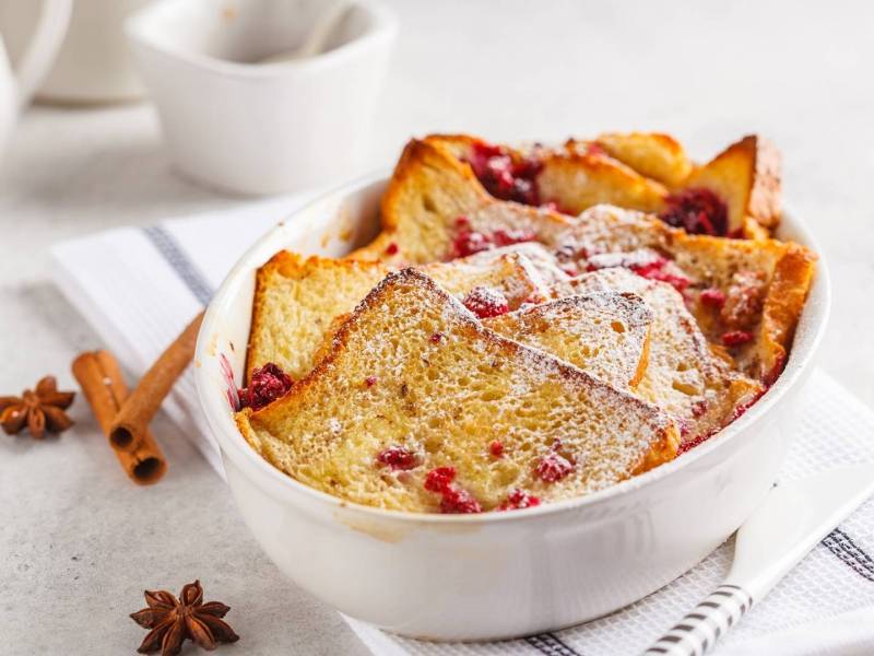 Toastbrotauflauf mit Himbeeren und Puderzucker in weißer Auflaufform, daneben Anissterne und Zimtstangen.