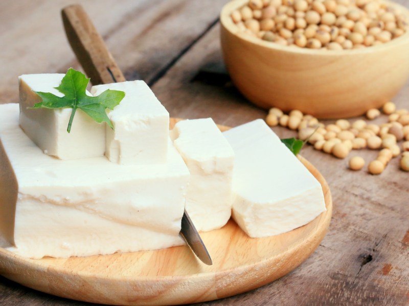 Ein Block Tofu und mehrere Stücke auf einem Holzbrett. Dahinter eine Schale mit Sojabohnen.
