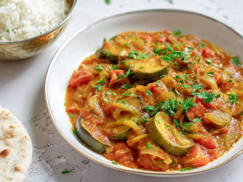 Teller mit einem Tomaten-Zucchini-Curry, Schale mit gekochtem Reis und Fladenbrot daneben