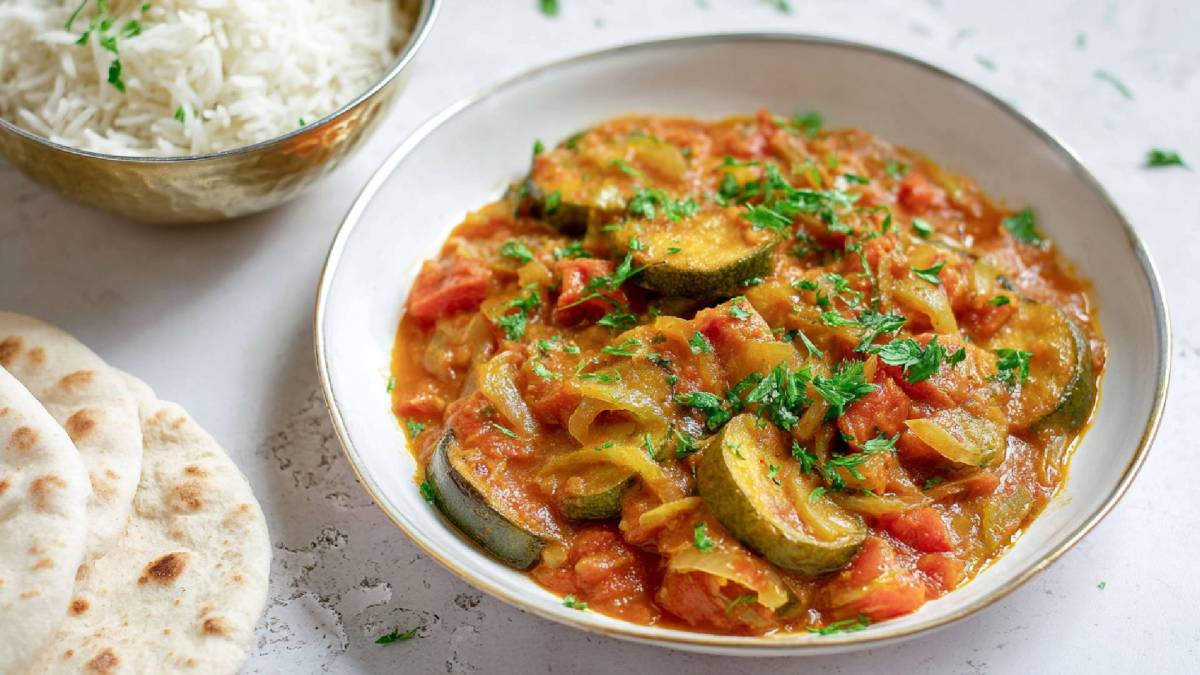 Teller mit einem Tomaten-Zucchini-Curry, Schale mit gekochtem Reis und Fladenbrot daneben