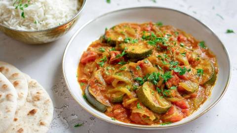 Teller mit einem Tomaten-Zucchini-Curry, Schale mit gekochtem Reis und Fladenbrot daneben