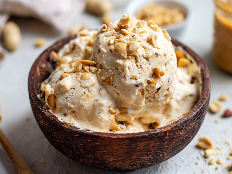 Eine kleine Holzschüssel, die mit einigen Kugeln veganem Erdnussbutter-Eis aus Seidentofu gefüllt ist und mit gehackten Erdnüssen garniert ist. Daneben liegt ein Löffel mit Erdnussbutter. Im Hintergrund steht ein Glas mit Erdnussbutter. Es liegen gehackte Erdnüsse und ganze Erdnüsse in der Schale herum.