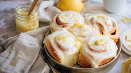Eine runde Auflaufform aus Keramik, die mit Zitronenschnecken gefüllt ist. Die Zitronenschnecken sind mit einem Frischkäse-Frosting getoppt und mit frischer Zitronenzeste garniert. Neben der Form ist ein Küchenhandtuch drapiert. Im Hintergrund ist ein Glas mit Lemon Curd sowie eine frische Zitrone zu sehen.