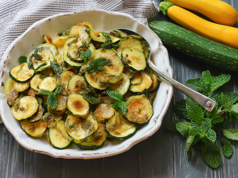 Ein Teller Zucchini alla Scapece, daneben Minzblätter und gelbe und grüne Zucchini.