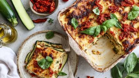 Draufsicht auf eine Auflaufform mit Zucchini-Feta-Lasagne. Ein Stück davon liegt auf einem Teller daneben. Ringsum sind frische Zutaten verteilt wie Zucchini, eine Schale mit getrockneten Tomaten, eine Glasgefäß mit Olivenöl, frisches Basilikum.