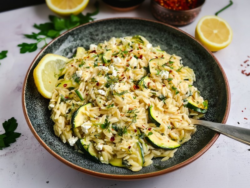Ein großer tiefer Teller mit Zucchini-Feta-Orzo, der mit frischer Petersilie und Chiliflocken garniert ist. Außerdem liegt eine Zitronenscheibe dabei. Um den Teller herum sind Zutaten zu sehen wie halbe Zitronen, frische Petersilie und eine Schale mit Chiliflocken.