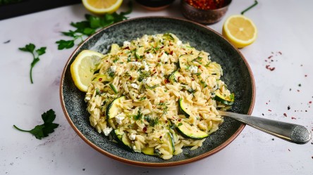 Ein großer tiefer Teller mit Zucchini-Feta-Orzo, der mit frischer Petersilie und Chiliflocken garniert ist. Außerdem liegt eine Zitronenscheibe dabei. Um den Teller herum sind Zutaten zu sehen wie halbe Zitronen, frische Petersilie und eine Schale mit Chiliflocken.