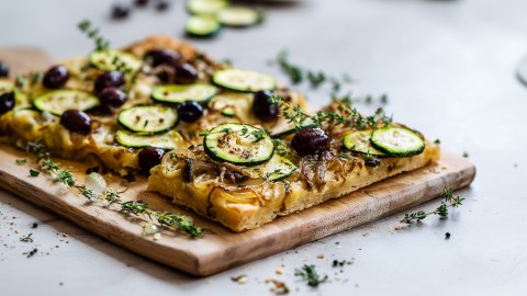 Eine Zucchini-Pissaladière, mit Zucchini, karamellisierten Zwiebeln, schwarzen Oliven und Sardellen. Die Pissaladière liegt auf einem Holzbrett. Ringsum liegt frischer Thymian verstreut.