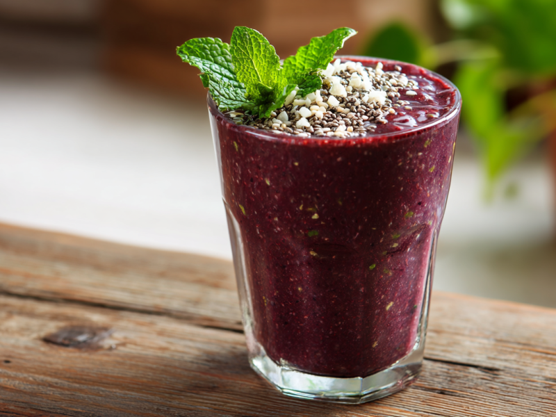 Ein Açaí-Smoothie im Glas steht auf einem Holztisch. Er ist mit Minze, Kokosraspeln und Chia-Samen garniert. Im Hintergrund sieht man noch eine Pflanze stehe.