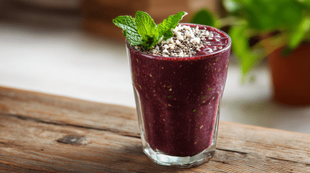 Ein Açaí-Smoothie im Glas steht auf einem Holztisch. Er ist mit Minze, Kokosraspeln und Chia-Samen garniert. Im Hintergrund sieht man noch eine Pflanze stehe.